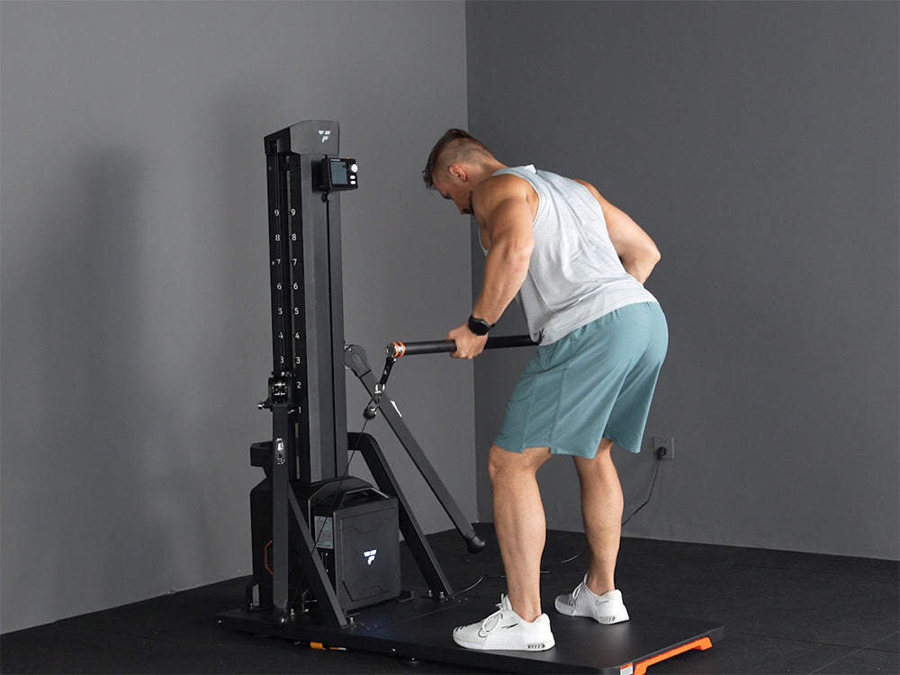 A man in a bent-over position using a bar attachment on a black digital functional trainer machine in a gym corner