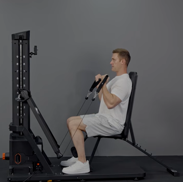 Man in white t-shirt and light gray shorts doing seated cable bicep curl on all-in-one home gym machine against plain gray wall