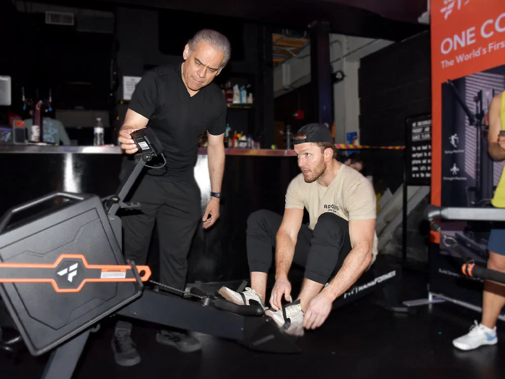 Senior fitness coach in black outfit adjusting fitness equipment parameter while young man in beige tee fixes foot strap on rowing-style gym machine at gym event venue