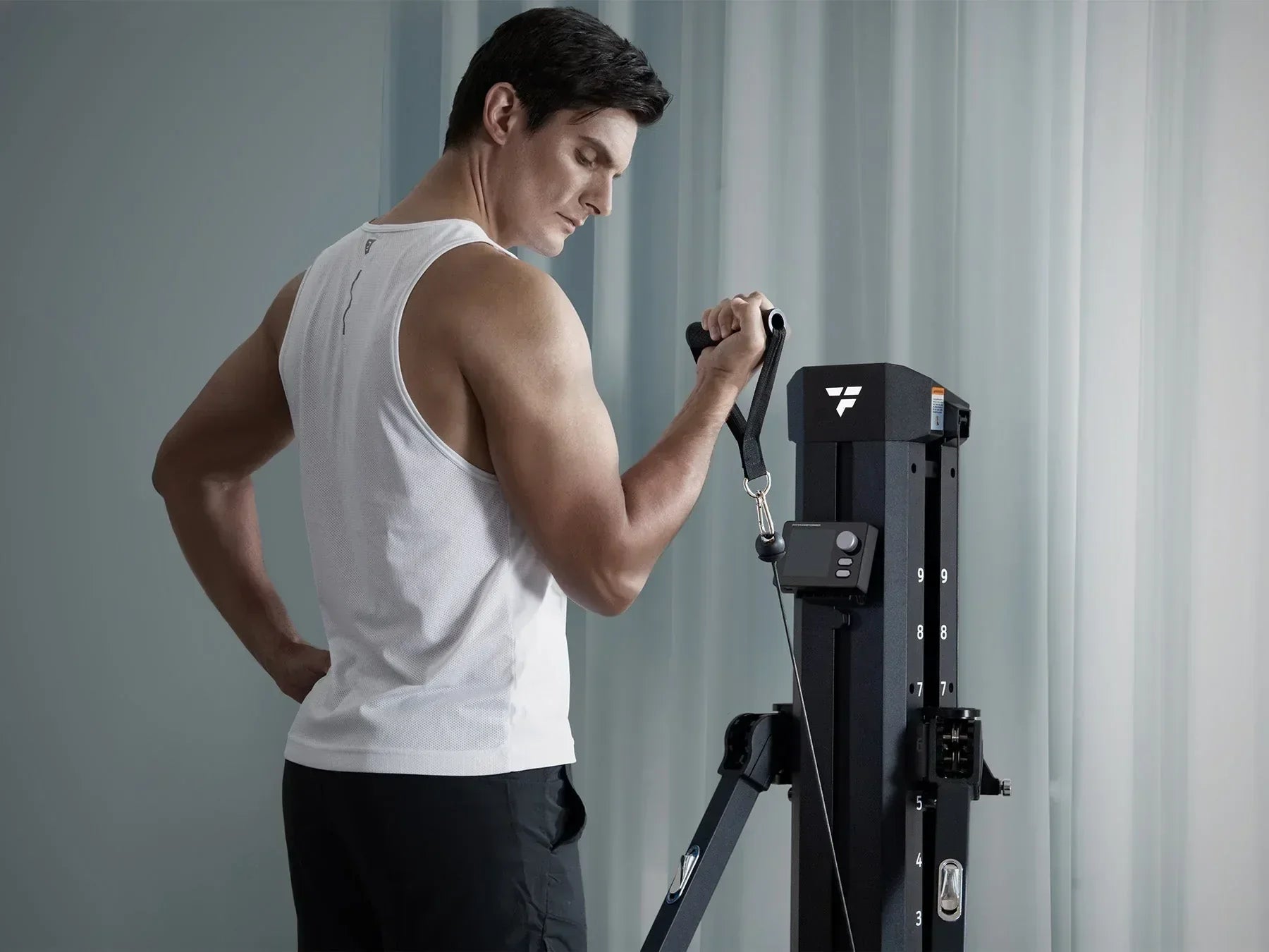 Athletic male in white sleeveless tank top and black pants doing cable bicep curl workout on vertical multifunctional home gym machine, soft grey curtain background