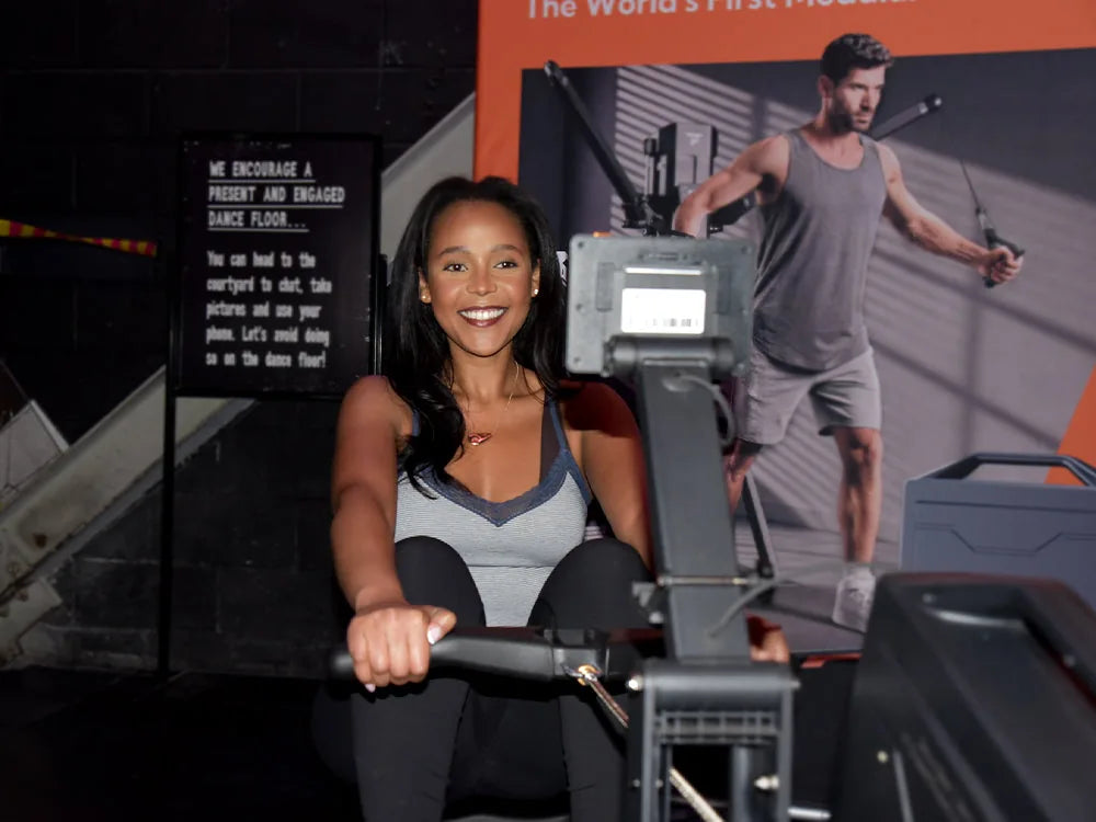 Smiling Black woman in grey lace-trim camisole and black leggings sits and rows on multifunctional fitness machine at indoor fitness event, orange promotional backdrop and black notice sign behind her