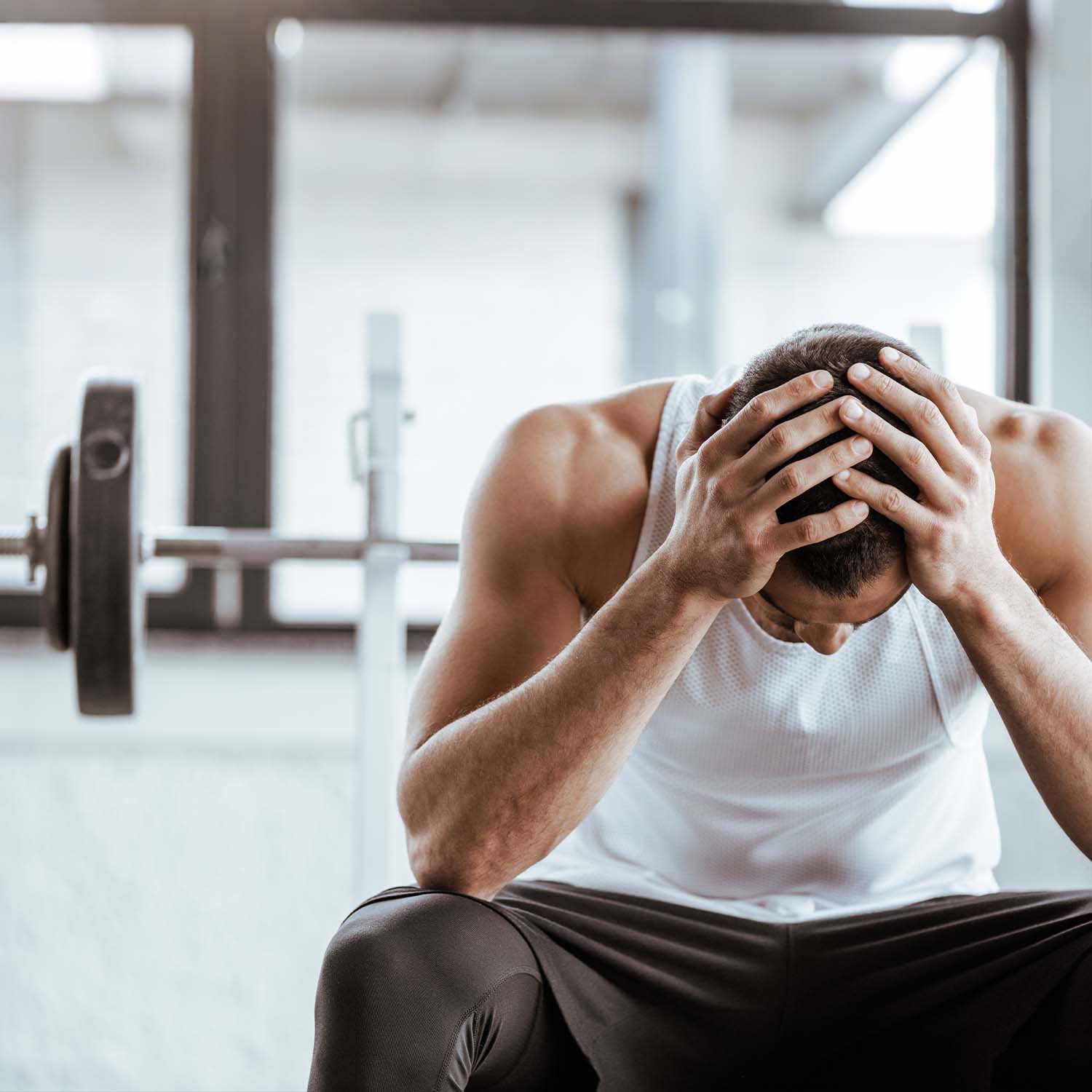 Frustrated man sitting on gym bench holding head showing workout burnout and overtraining symptoms in fitness center